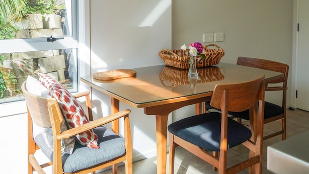 A dining area with a glass table, wooden chairs, a wicker basket, and a vase with flowers. - Private, spacious, well appointed, self-contained unit. (Tauranga)