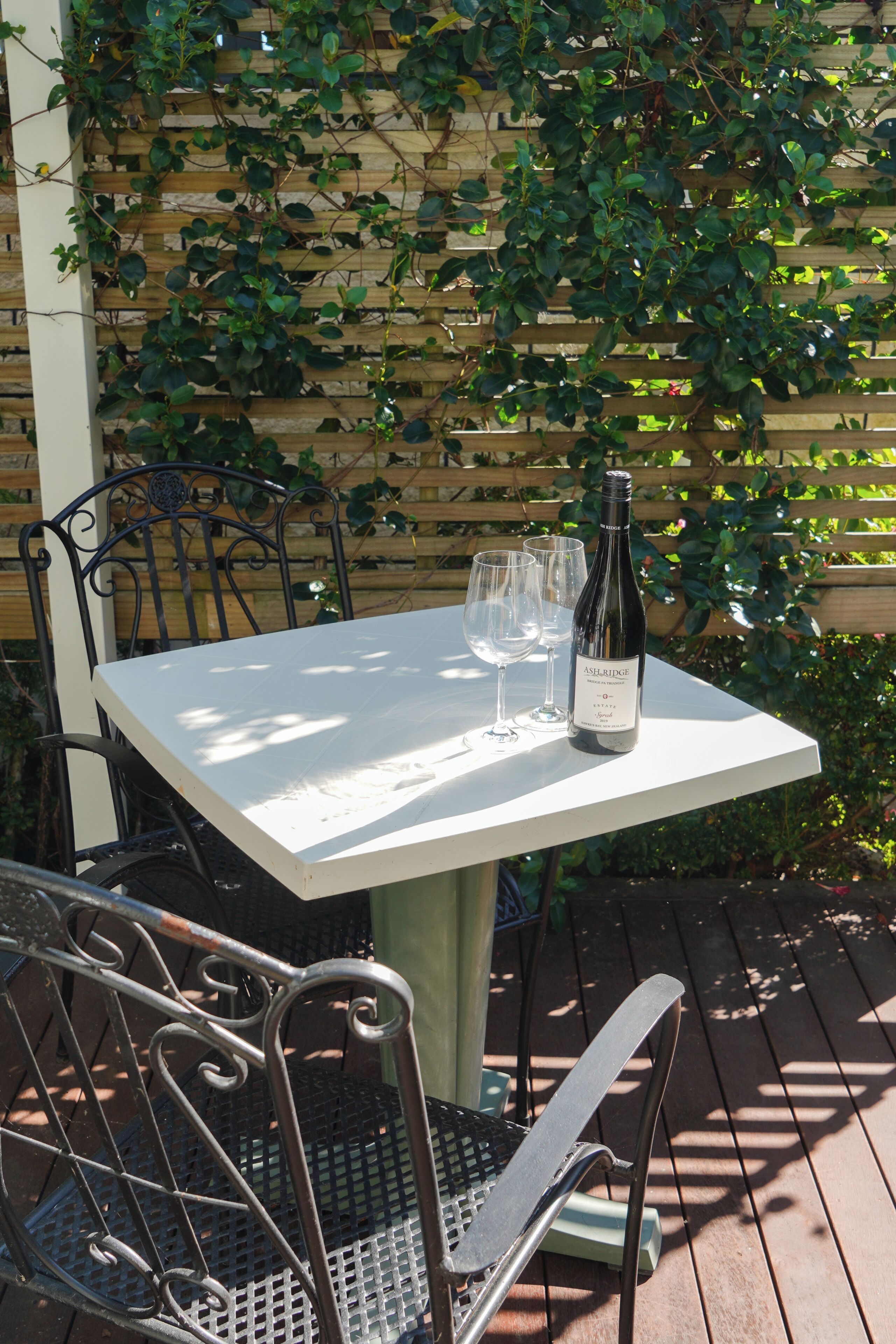 Une table avec une bouteille et des verres, entourée de chaises en métal, installée à l’extérieur, avec une clôture en bois et de la verdure en arrière-plan.