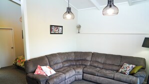 A corner living room with a brown sectional sofa, two pendant lights, a framed artwork, and a small fan on a shelf. - Private, spacious, well appointed, self-contained unit. (Tauranga)
