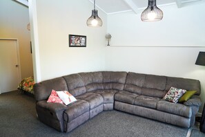 Ein Eckwohnzimmer mit einem braunen Modulsofa, zwei Pendelleuchten, einem gerahmten Kunstwerk und einem kleinen Ventilator auf einem Regal.