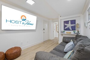 A living room with a grey sofa, two brown ottomans, a mounted TV displaying 'HOST & HOME Luxury Vacation Rentals', and a hallway leading to another room. - 2BR Villa w/ Bonus Lounge & Wine Fridge, Indoor & Outdoor Pools, Near Coligny (Hilton Head Island)