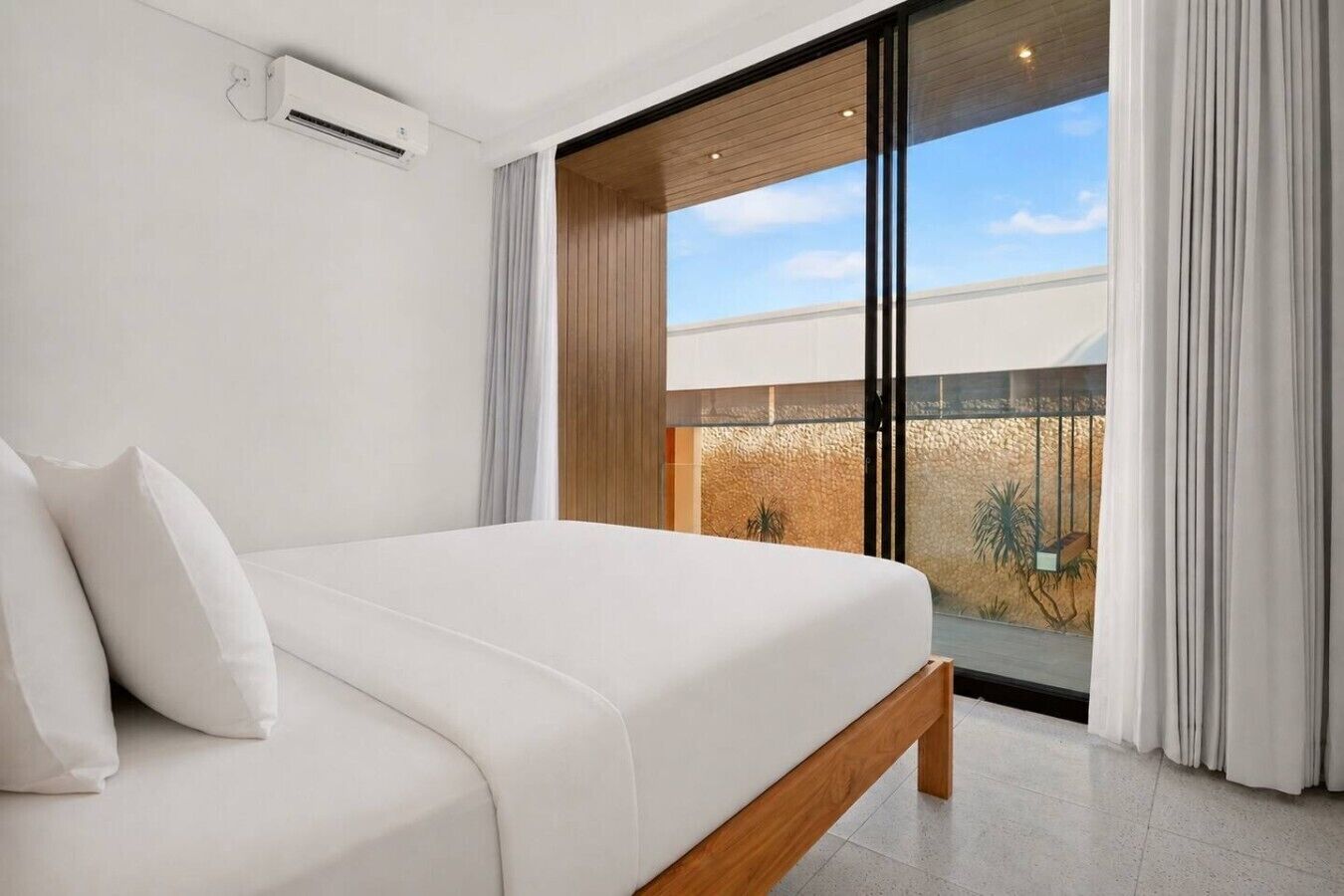 Une chambre moderne avec un grand lit, une porte-fenêtre coulissante donnant sur un balcon et un espace extérieur visible, agrémenté de plantes.