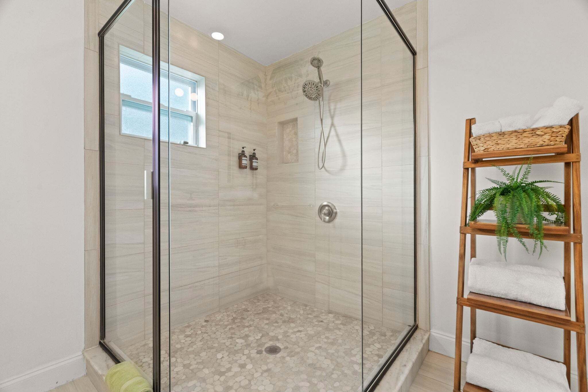 A glass-enclosed shower with a tiled floor, a wooden shelf with towels, and a window.