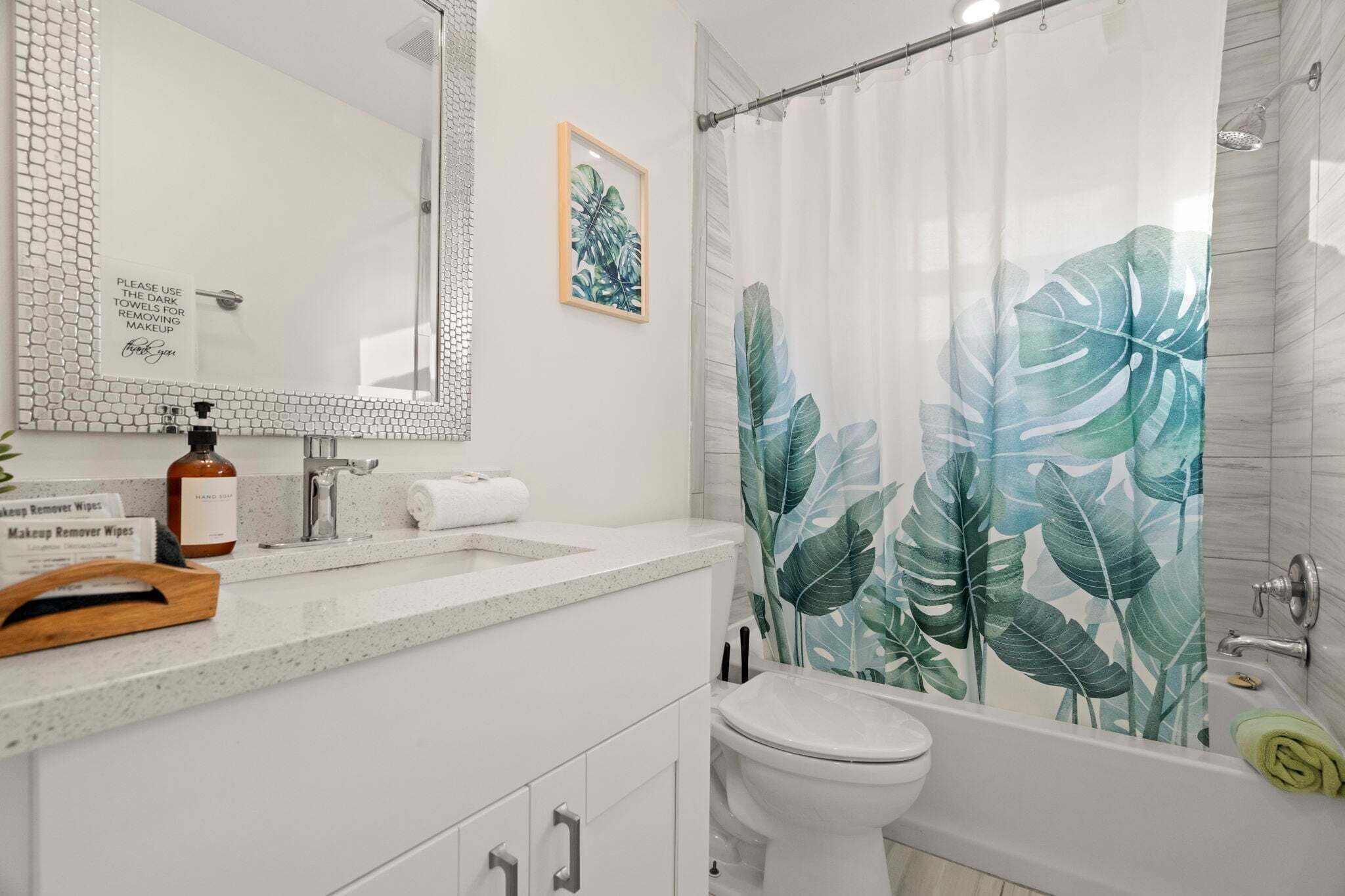 Une salle de bain avec un lavabo blanc, un miroir et des toilettes. Il y a un rideau de douche orné d’un motif de feuilles tropicales.