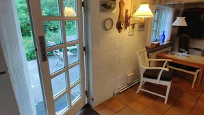 Une pièce avec un sol carrelé, une chaise blanche, une table à manger, une porte avec des panneaux de verre et une fenêtre donnant sur un paysage verdoyant.