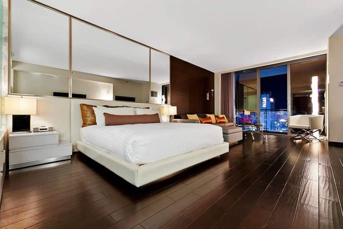 A modern hotel room with a large bed, a sofa, a desk, and a balcony with a city view.