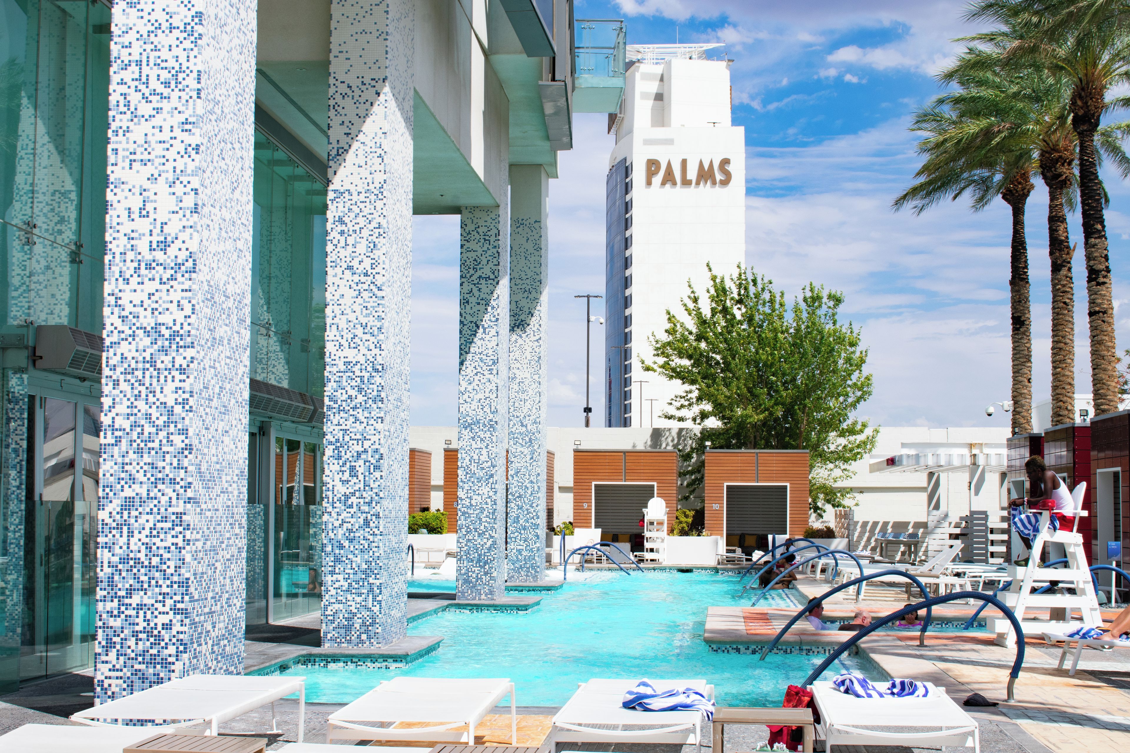 Een zwembadterras met betegelde zuilen, ligbedden en een gebouw met de aanduiding 'PALMS'.