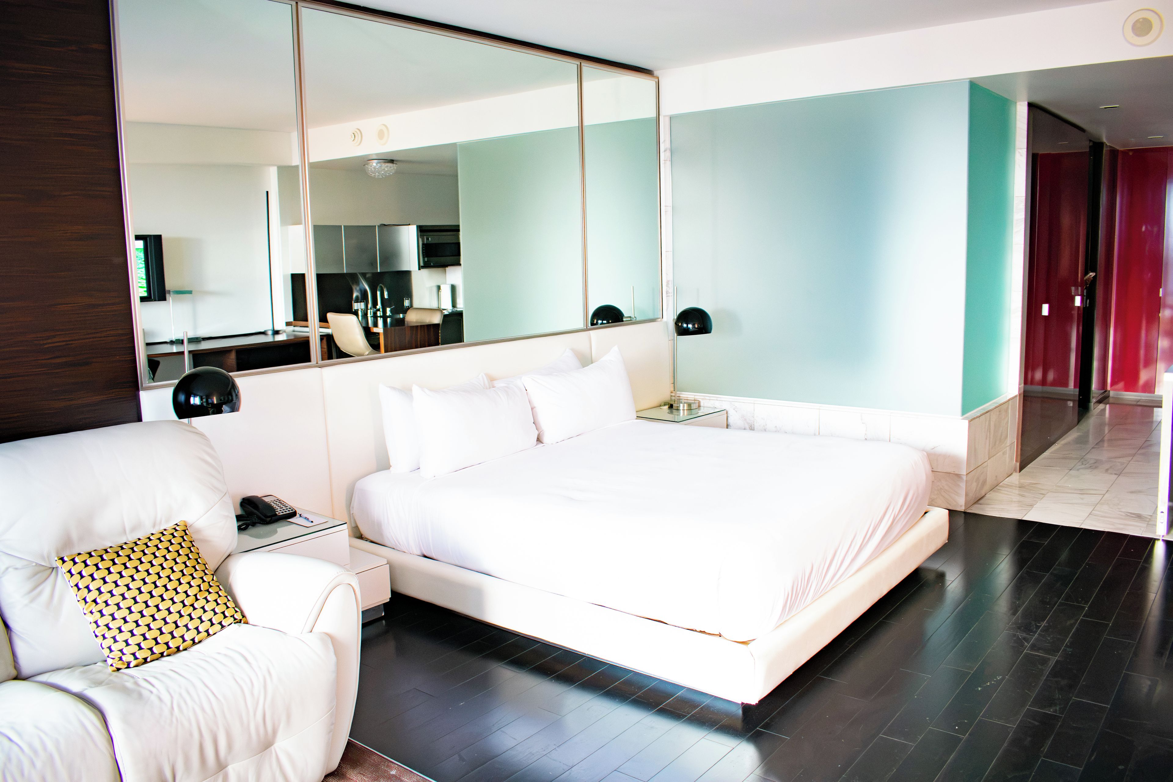 Une chambre d’hôtel moderne dotée d’un grand lit, d’un coin salon avec un fauteuil, d’une table de chevet, d’un miroir et donnant sur un couloir.
