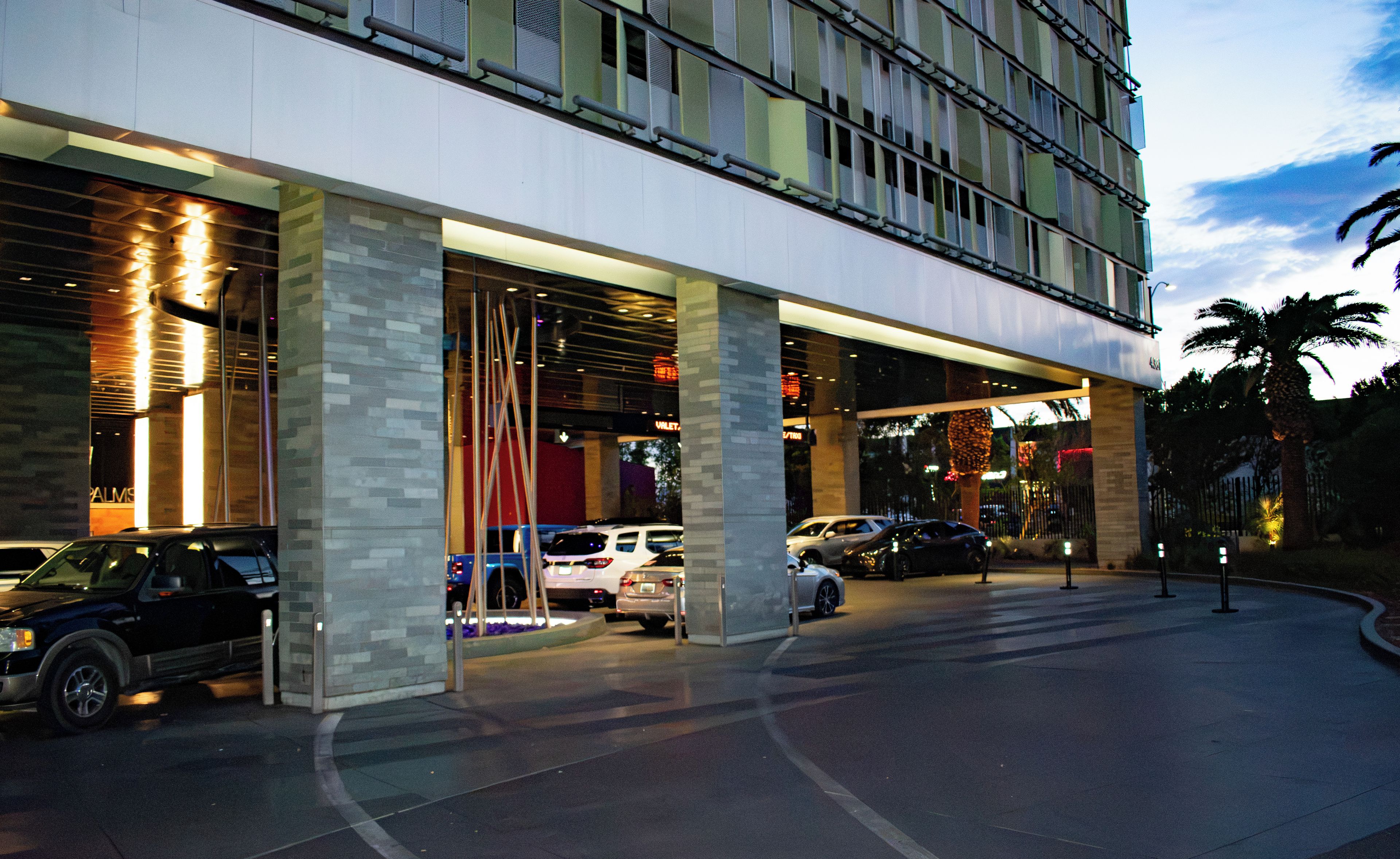 A hotel entrance with multiple cars parked in front, a modern building facade, and a palm tree in the background.