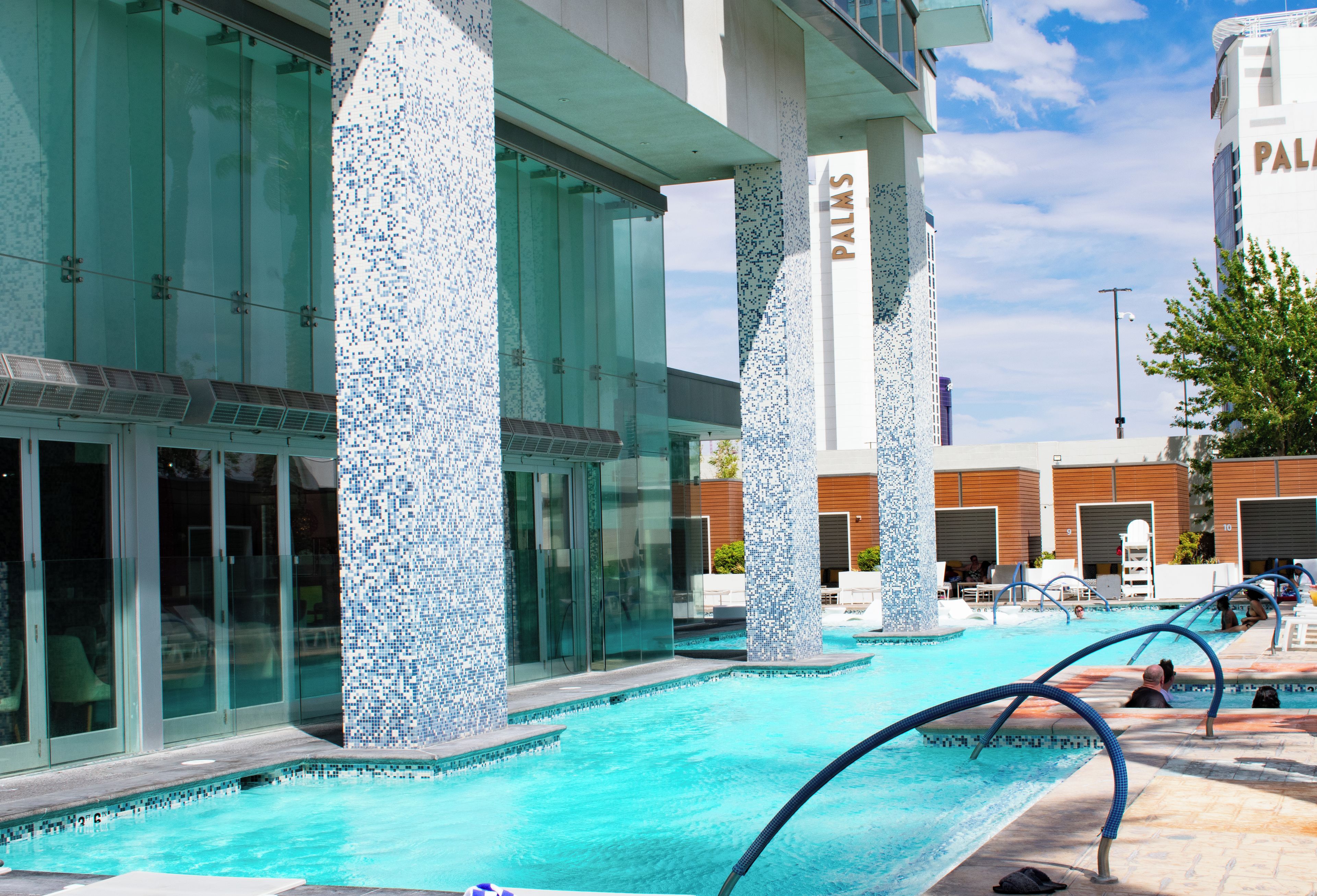 A swimming pool area with clear blue water, surrounded by glass walls and columns with mosaic tile patterns.