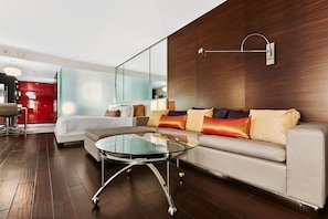 A modern hotel room with a glass coffee table, a sofa with cushions, a red accent wall, and a large mirror.