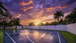 Ein Basketballplatz im Freien mit Sonnenuntergangsblick, umgeben von Palmen und einer weißen Mauer.