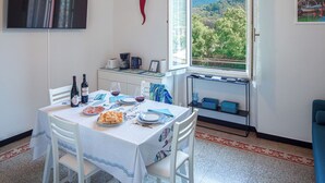 Zona pranzo con tavolo apparecchiato per due, una finestra vista alberi e colline, e angolo cottura con macchina del caffè e tostapane.