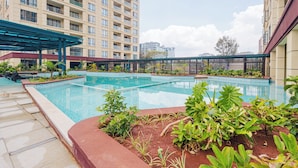 En swimmingpool omgiven av en trädgård, ett avskärmat sittområde och en hög bostadsbyggnad i bakgrunden.