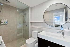 Un baño moderno con ducha envuelta en vidrio, inodoro blanco, lavabo blanco con espejo y repisa con artículos de aseo.