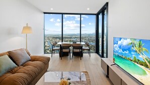 Ein modernes Wohnzimmer mit großem Fernseher, einer Couch, einem Esstisch mit Stühlen und Blick auf eine Stadtlandschaft durch große Fenster.