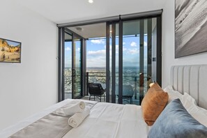 A bedroom with a large bed, a view of a cityscape through a sliding glass door, and a painting on the wall. - Stylish 1BR Apartment in Broadbeach (Broadbeach)