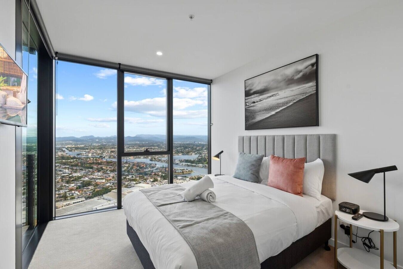 A modern bedroom with a large bed, a city view, a framed picture, and a bedside table with a lamp.