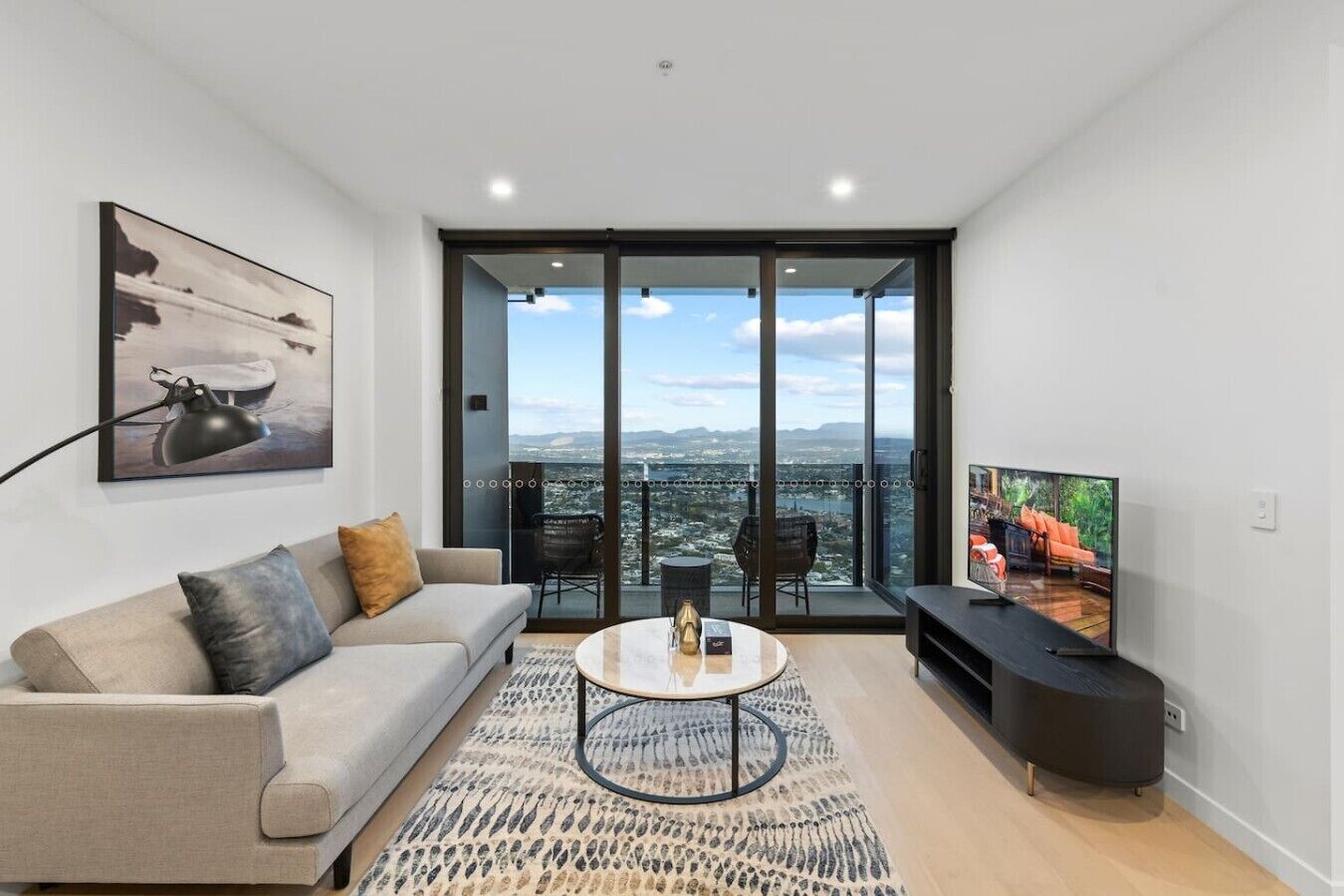 Ein modernes Wohnzimmer mit Sofa, Couchtisch und Fernseher, mit Blick auf die Stadt durch große Glasfenster.