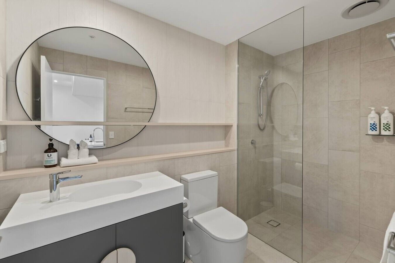 A modern bathroom with a glass-enclosed shower, a round mirror, and a white sink.