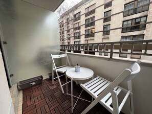 Ein Balkon mit weißem Stuhl, einem kleinen runden Tisch und Blick auf ein Gebäude mit vielen Fenstern.