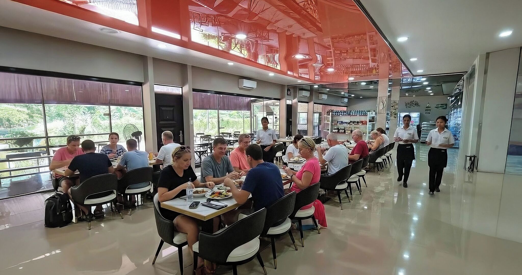 A spacious dining area with multiple tables and chairs, large windows offering views of greenery, and a modern ceiling design.