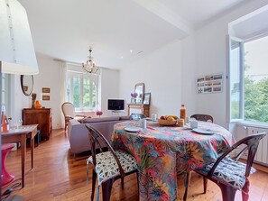 A dining area with a round table, chairs, and a colorful tablecloth. There is a television, a wooden cabinet, and a window with a view of greenery. - Ty Yannick by Interhome (Ploumanach)