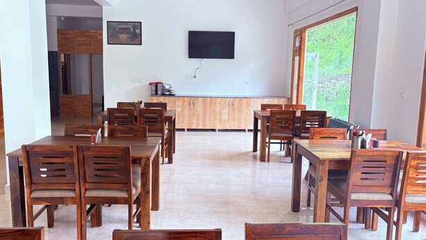 A dining area with wooden tables and chairs, a television mounted on the wall, and a large window providing a view of greenery. - The Kath Kuni (Kullu)