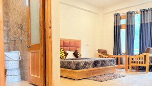 A bedroom with a bed, wooden headboard, two chairs, a small table, and a bathroom with a shower and toilet. - The Kath Kuni (Kullu)