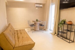 Una sala de estar con un sofá, una mesa de comedor con sillas, un televisor montado en la pared y una planta en maceta.