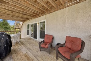 Un patio cubierto con suelo de madera, dos sillas de mimbre con cojines, una parrilla y una puerta corrediza de vidrio.