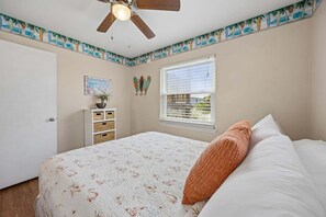 Un dormitorio con una cama, un ventilador de techo, una ventana con persianas, una puerta y una pared con un borde de temática tropical.