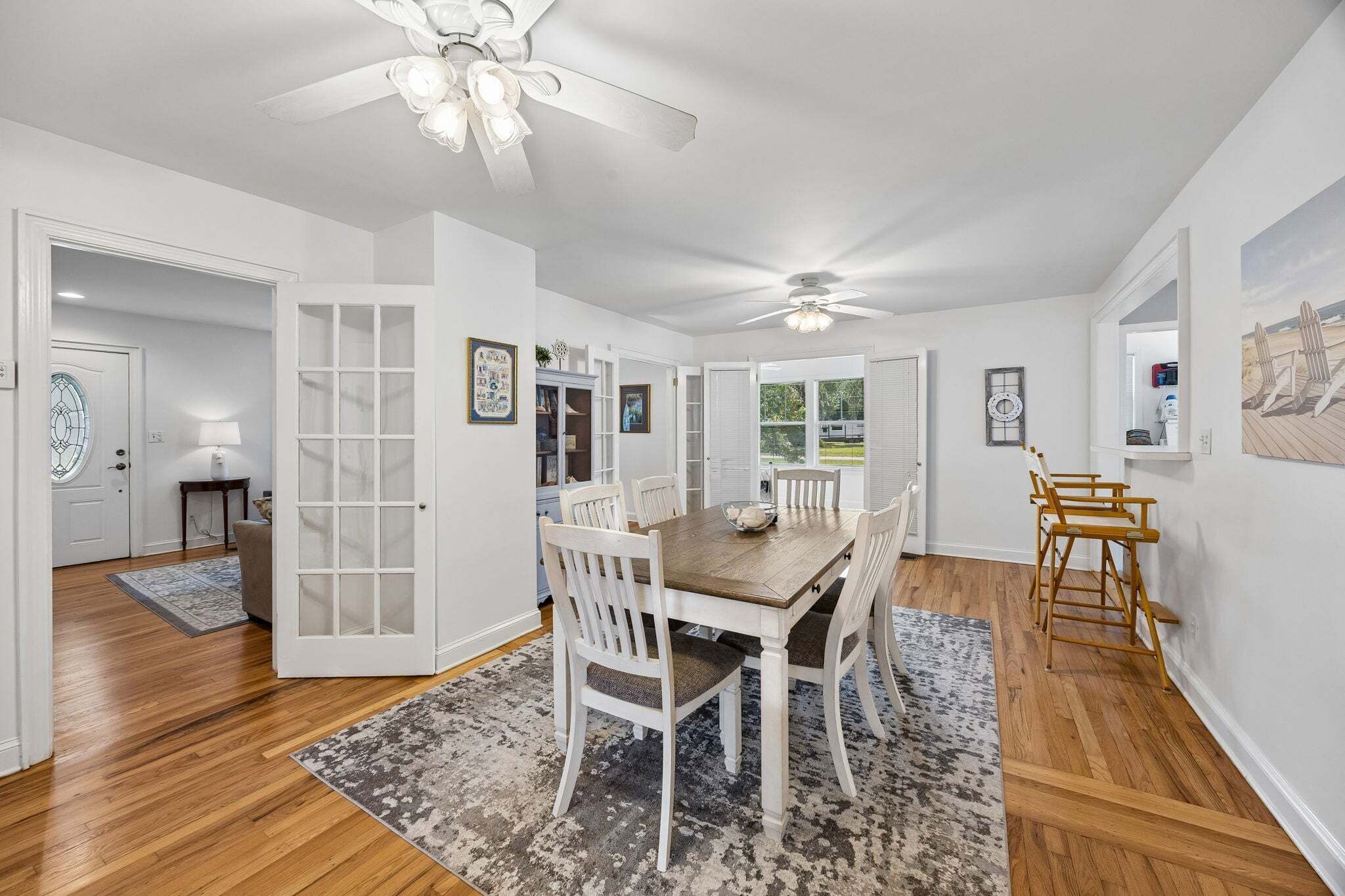 A dining area with a wooden table and white chairs, a ceiling fan, and a view of the outdoors.