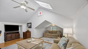 A spacious loft with a high ceiling, a ceiling fan, a large window, a flat-screen TV, a wooden entertainment unit, a beige sofa, a patterned throw pillow, a lamp, and a small table with a tray. - Downtown St. Marys Retreat | Sleeps 2 (St. Marys)