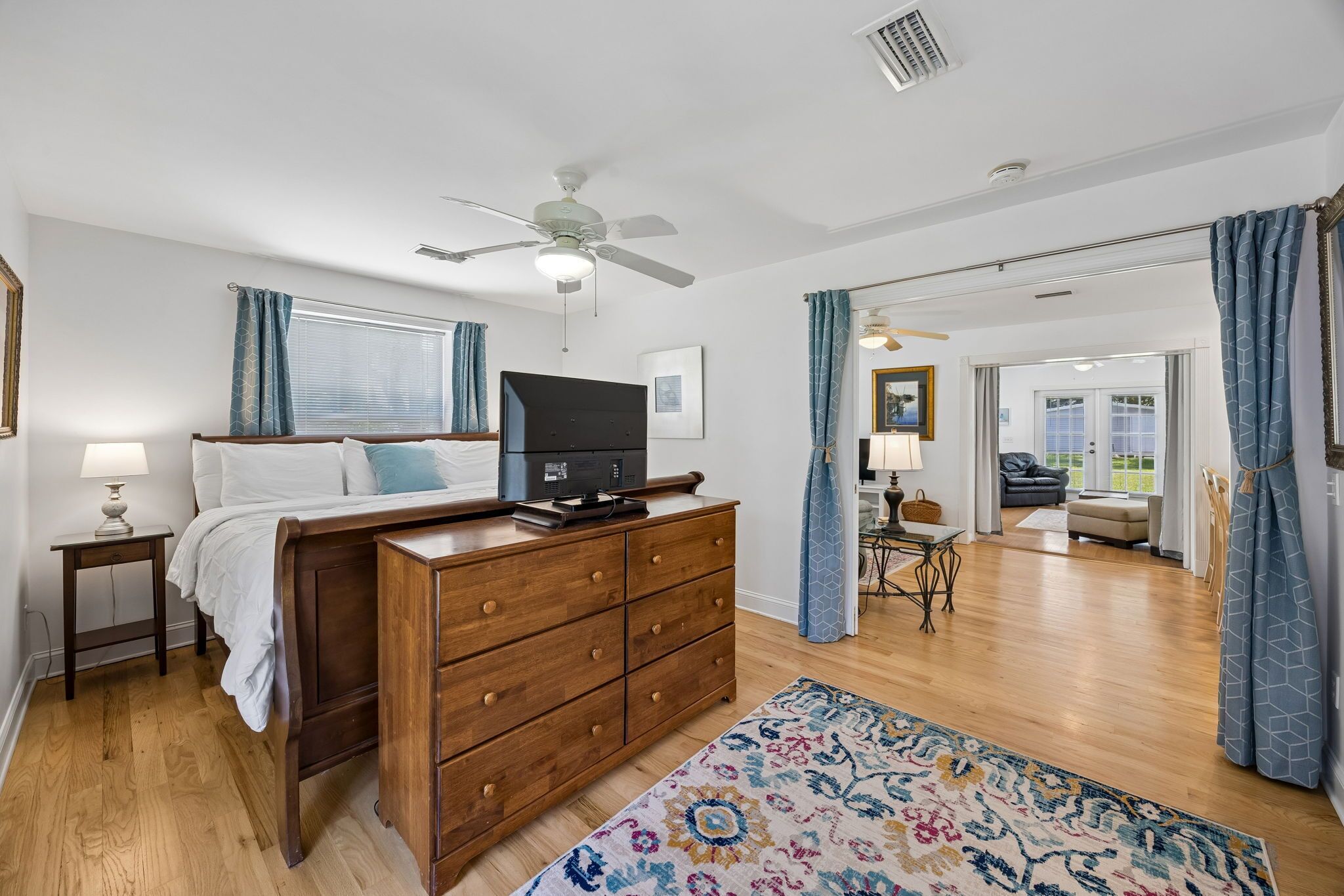 A bedroom with a wooden bed, a ceiling fan, a television, and a view to a living area.
