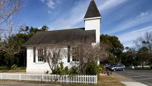Uma pequena igreja branca com um pináculo, rodeada por árvores e uma cerca branca de piquete.