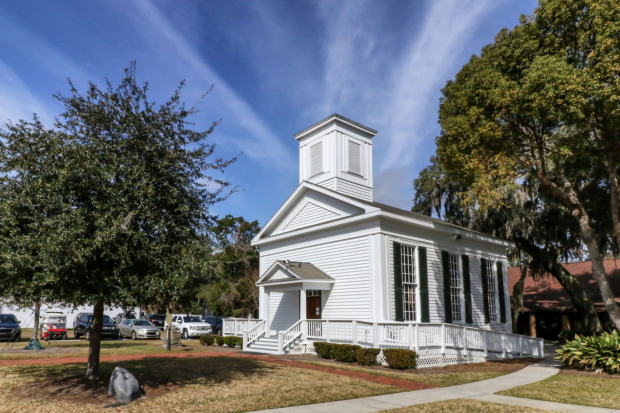 A white church with a steeple, surrounded by trees and parked cars.