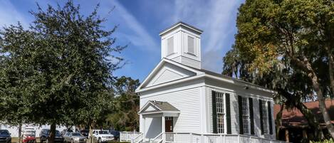 A white church with a steeple, surrounded by trees and parked cars.