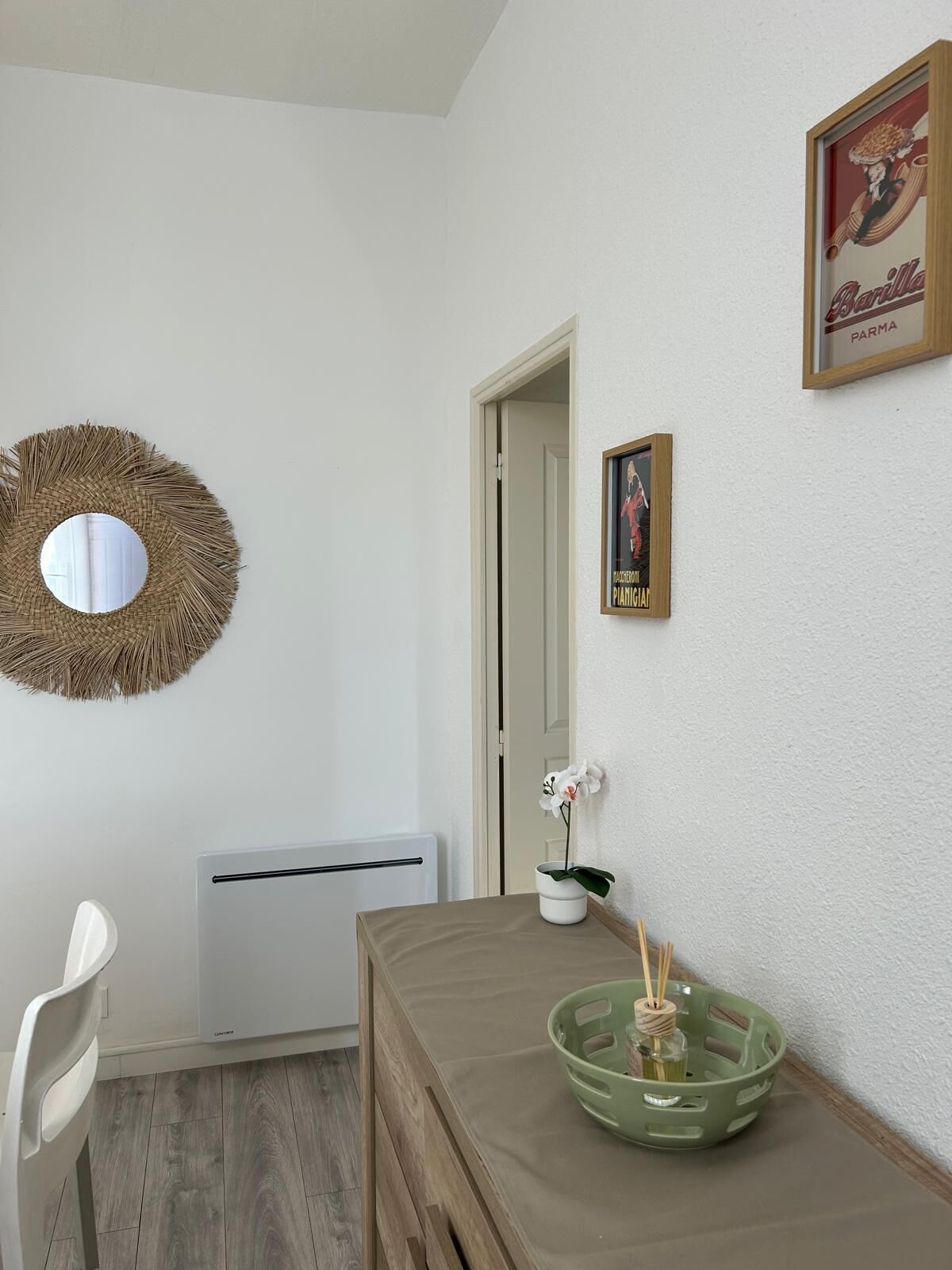 A room with a round mirror, a framed picture, a radiator, a table with a green tray, and a white chair.
