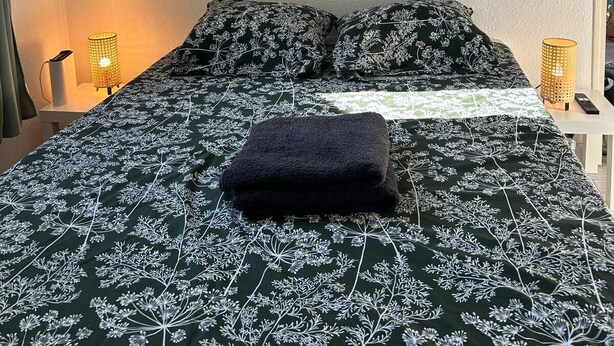 Una cama con colcha floral, dos almohadas, una manta y un cuadro de un paisaje costero en la pared.