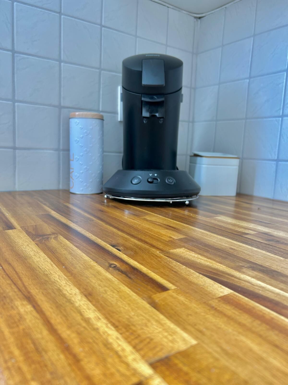 Eine Kaffeemaschine auf einem Holztisch vor einer Wand mit weißen Fliesen.