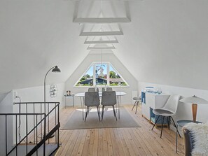 A modern loft bedroom with a sloped ceiling, a dining area with a round table and chairs, and a view of a park through a large window.
