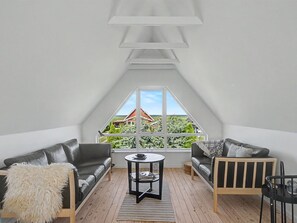 A loft bedroom with a sloped ceiling, two leather sofas, a small round table, and a large window offering a view of greenery and a distant building. - "Rosalie" - 150m from the sea by Interhome (Otterup)