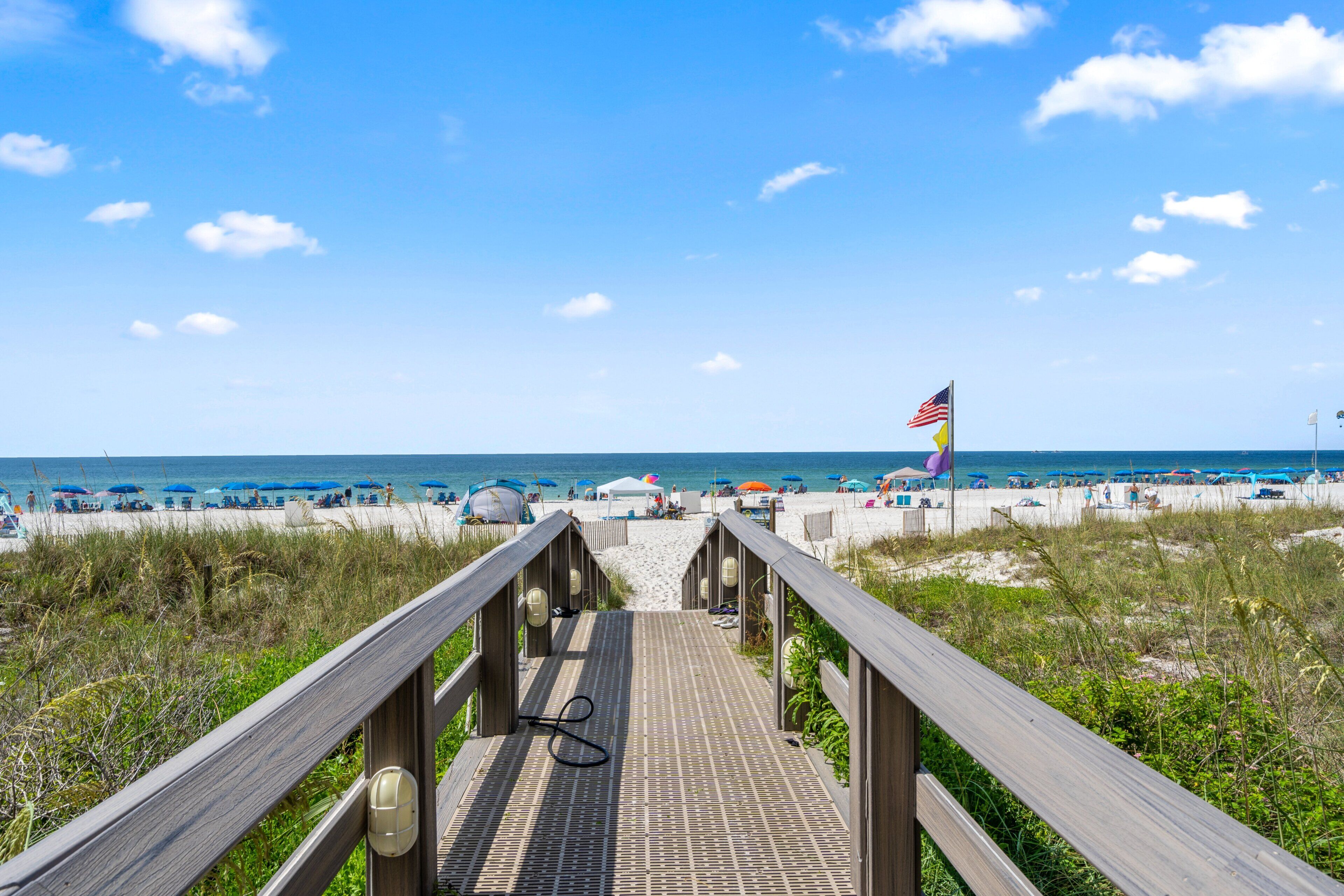 A wooden boardwalk leading to a beach with umbrellas and a flag.