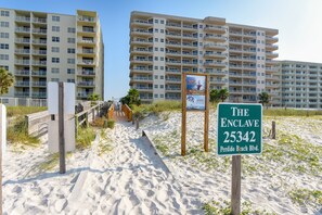 A beachside hotel named 'The Enclave' with a signboard, a sandy path, and a wooden fence. - The Enclave 801 | Gulf Front Corner Condo | Wraparound Balcony | My Beach Getaways (Orange Beach)