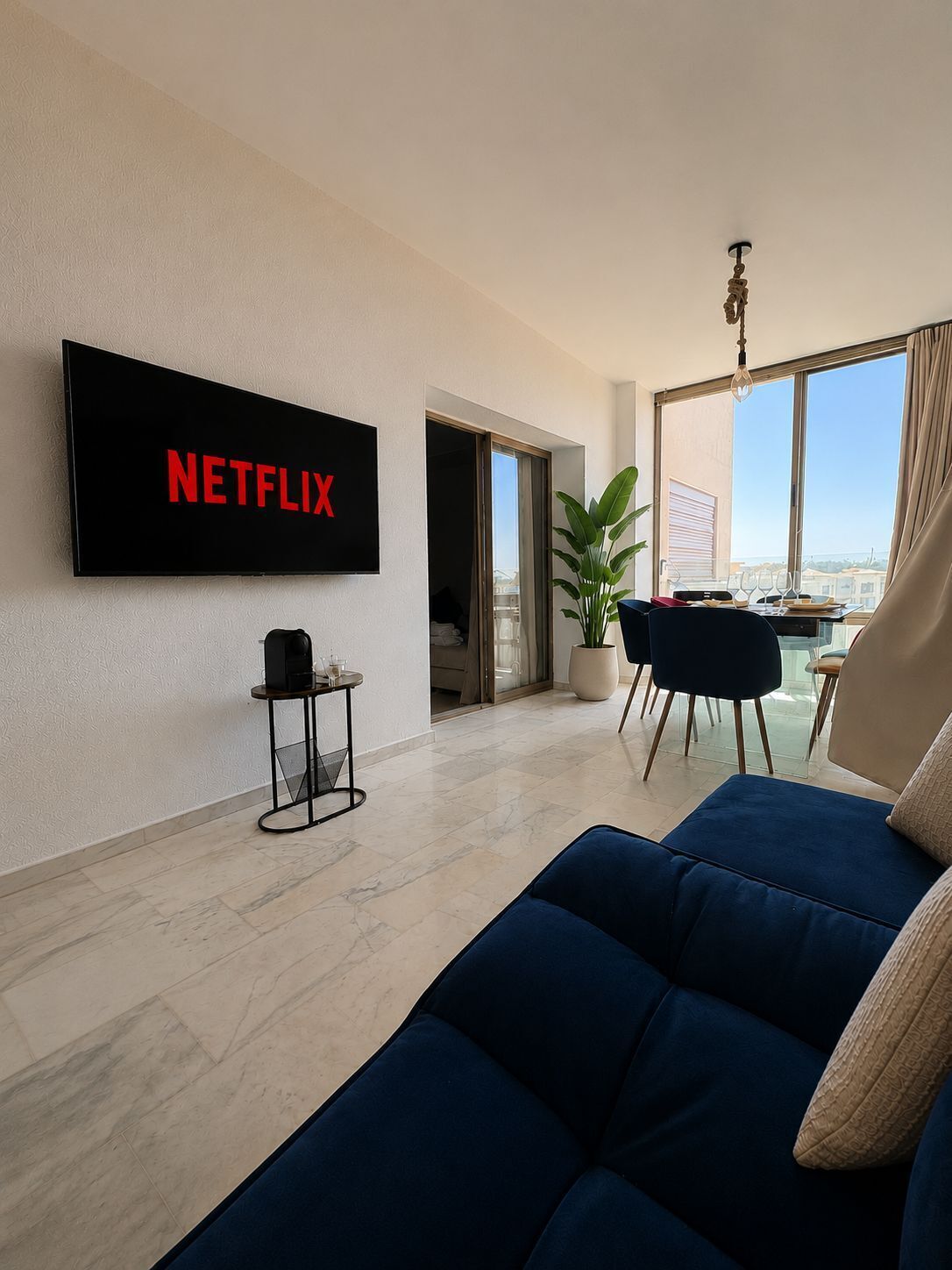 Ett modernt vardagsrum med en stor tv-skärm som visar ”NETFLIX”, en soffa, ett litet bord med en högtalare och en krukväxt.