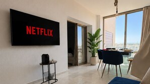 Un soggiorno moderno con una grande televisione a schermo piatto che mostra 'NETFLIX', un divano, un tavolino con un altoparlante e una pianta in vaso.