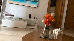 Un soggiorno con una TV a schermo piatto, un mobile TV bianco, un tavolino in legno con un vaso di fiori e un tappeto decorato.