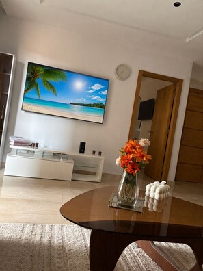 A living room with a flat-screen TV, a white entertainment unit, a wooden coffee table with a vase of flowers, and a patterned rug. - Apartment with a view of the pool in a beachfront residence (Bouznika)