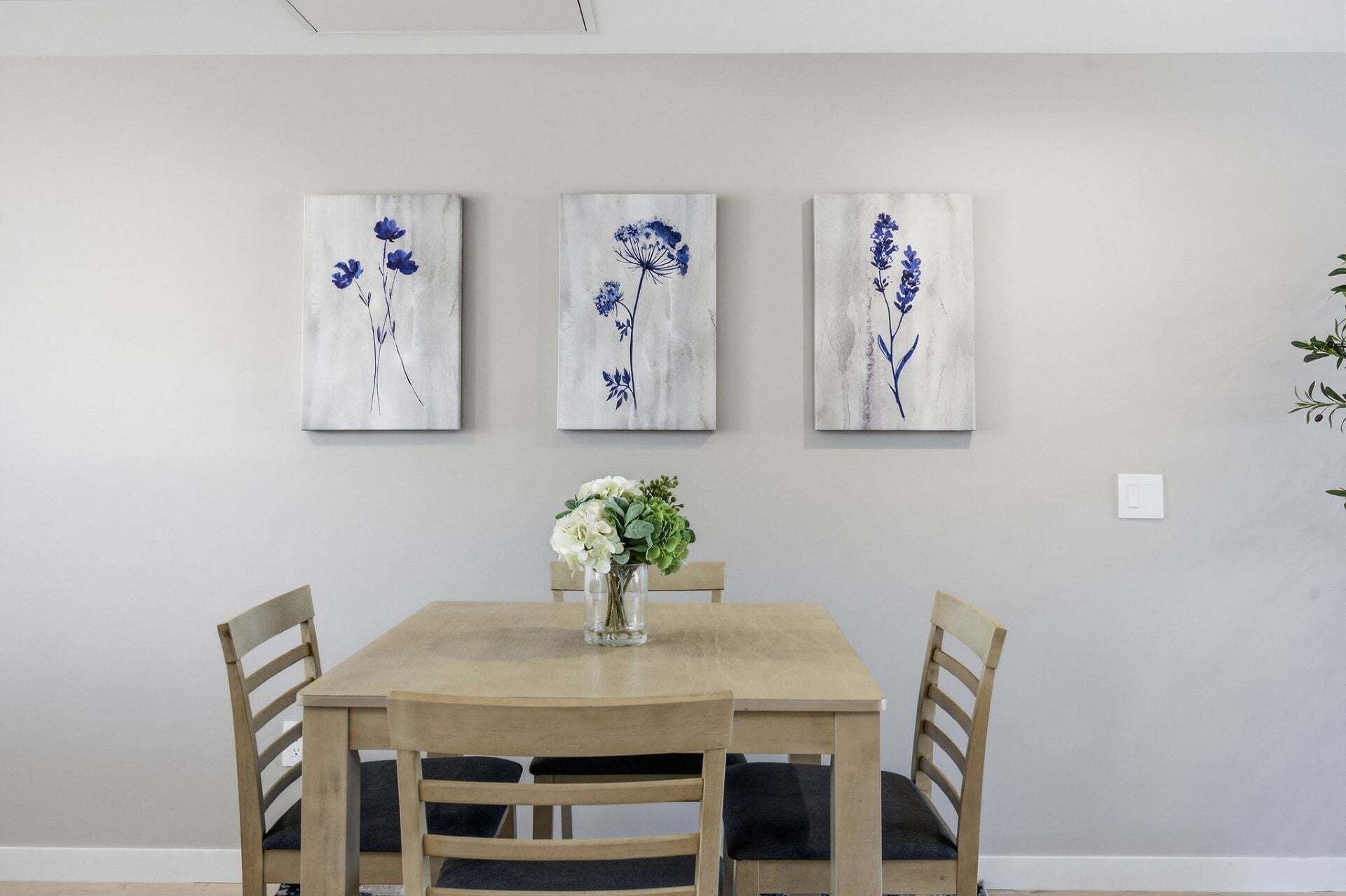 Un coin repas avec une table et des chaises en bois, un vase rempli de fleurs et trois tableaux représentant des fleurs bleues, accrochés au mur.